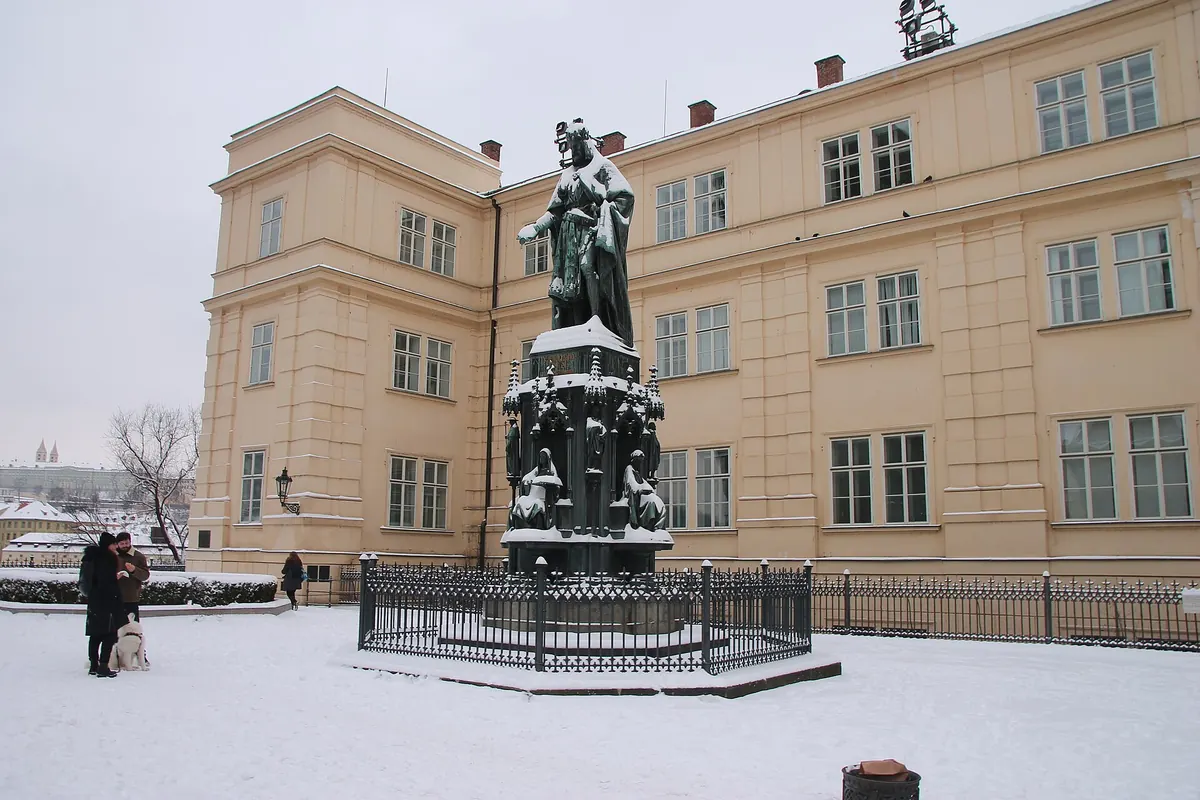 En statue af Karl IV på en stenplint på en sneklædt plads, med en lang lysegul bygning bagved